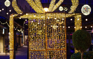Light up gold present box displayed outdoors with warm white aurora balls and festoon lights suspended above it at Woolshops Shopping Centre.