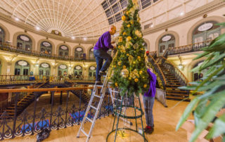 Two members of Fizzco's installation team installing an artificial cone Christmas tree decorated with gold baubles at Leeds Corn Exchange.