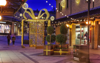 Light up gold present box displayed outdoors with warm white aurora balls and festoon lights suspended above it at Woolshops Shopping Centre.