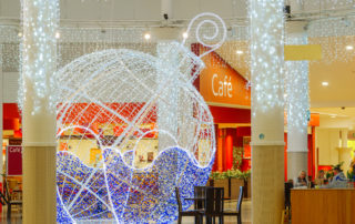 Light up bauble motif displayed in the centre of a shopping centre with bright white curtain lights around the pillars and the roof in the shopping centre.