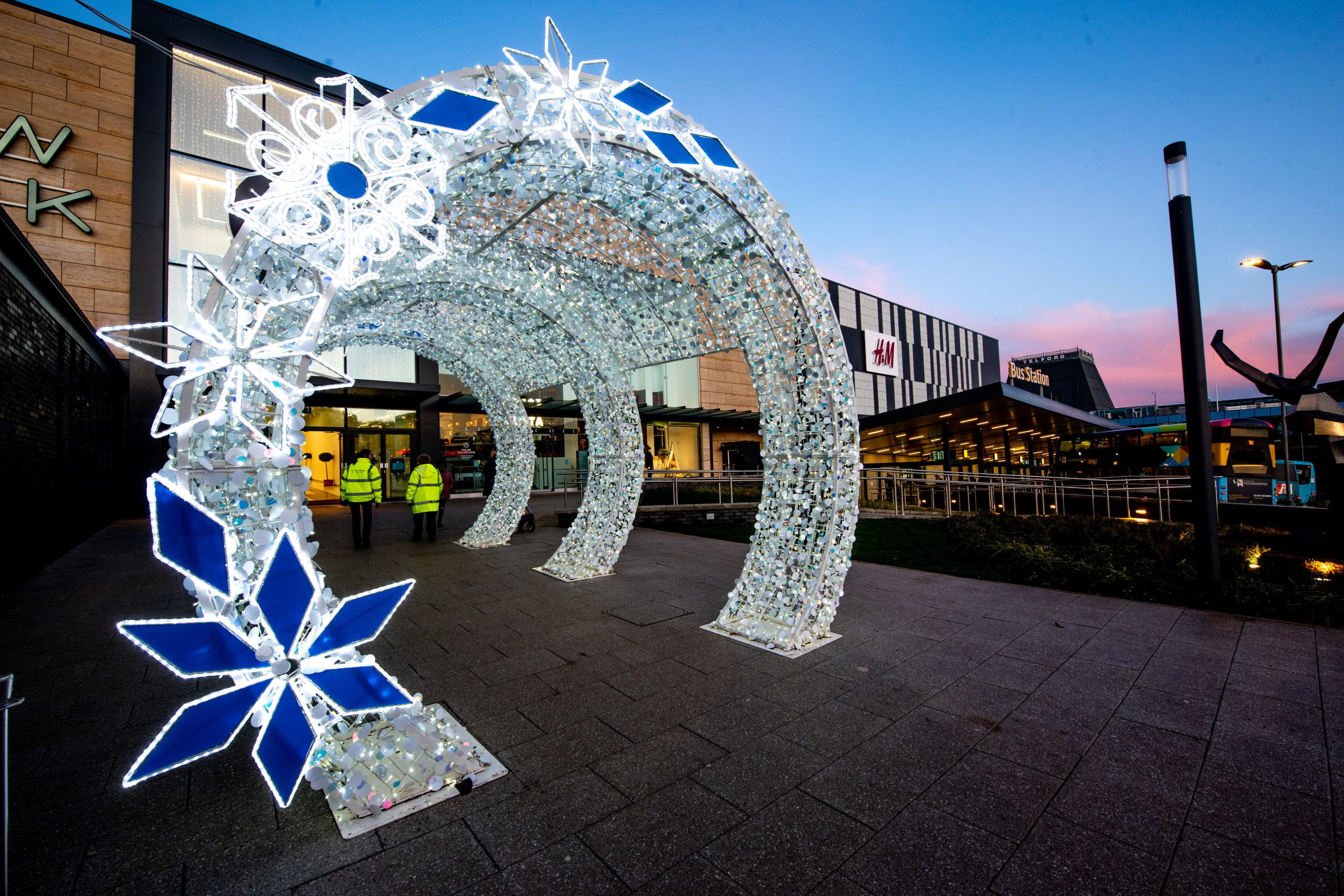 Bright white light up cylinder walkthrough tunnel decorated with silver tinsel and white and blue snowflakes leading to the entrance of Telford Shopping Centre.