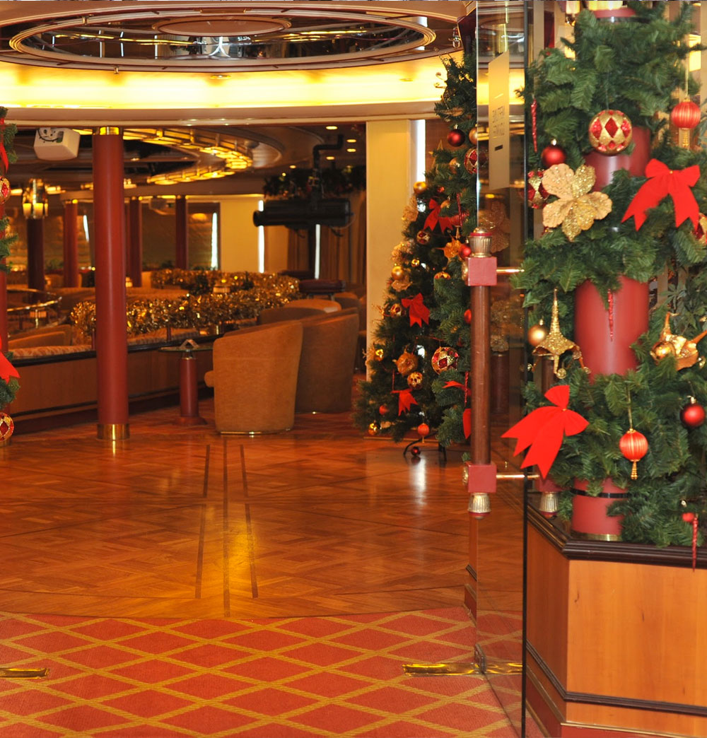 Cruise ship Christmas display featuring an artificial green Christmas tree and a green garland wrapped around a pillar, both decorated with traditional red and gold baubles, bows and flowers.