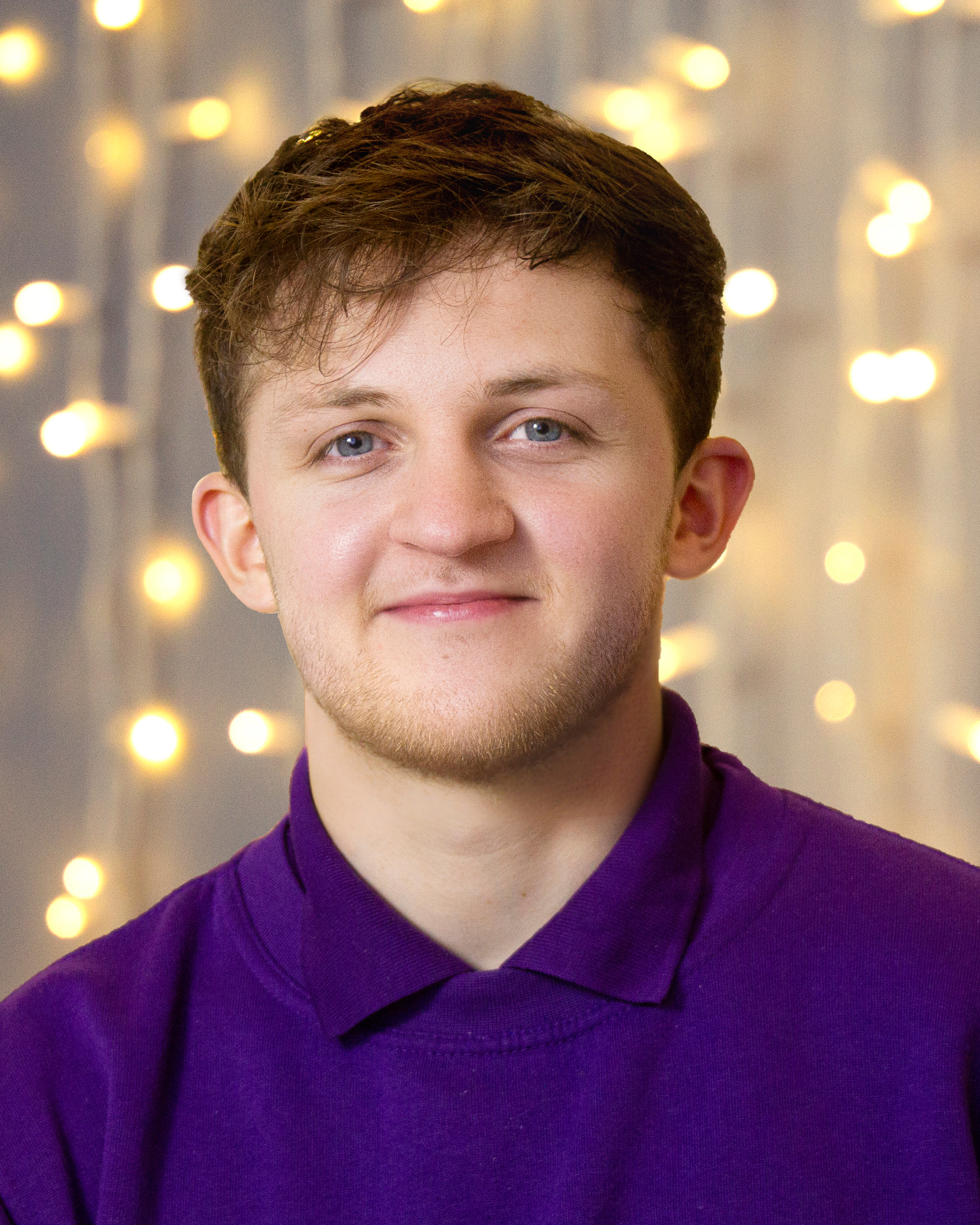 A headshot image of a Fizzco employee wearing Fizzco’s purple uniform in front of warm white curtain lights.