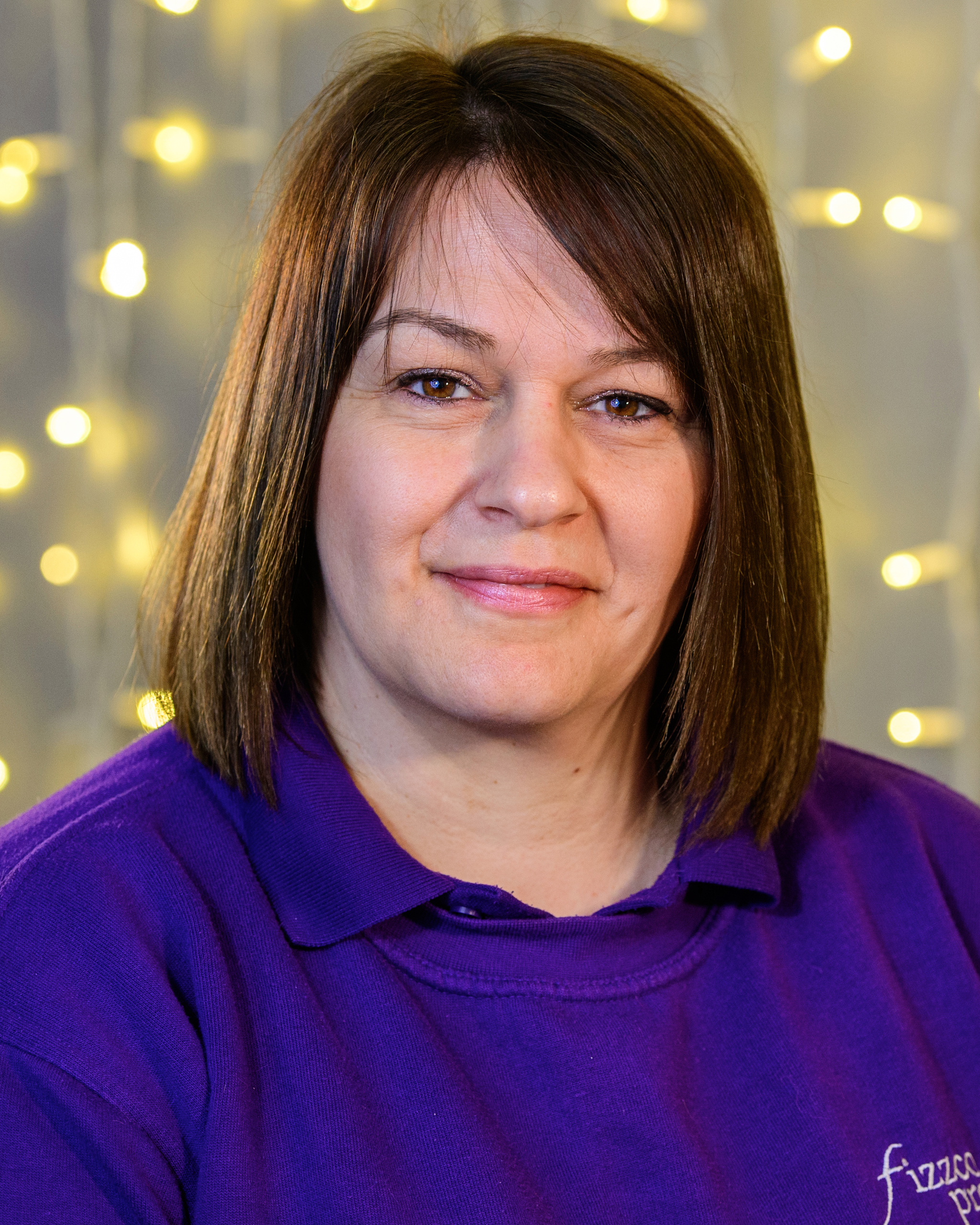 A headshot image of a Fizzco employee wearing Fizzco’s purple uniform in front of warm white curtain lights.