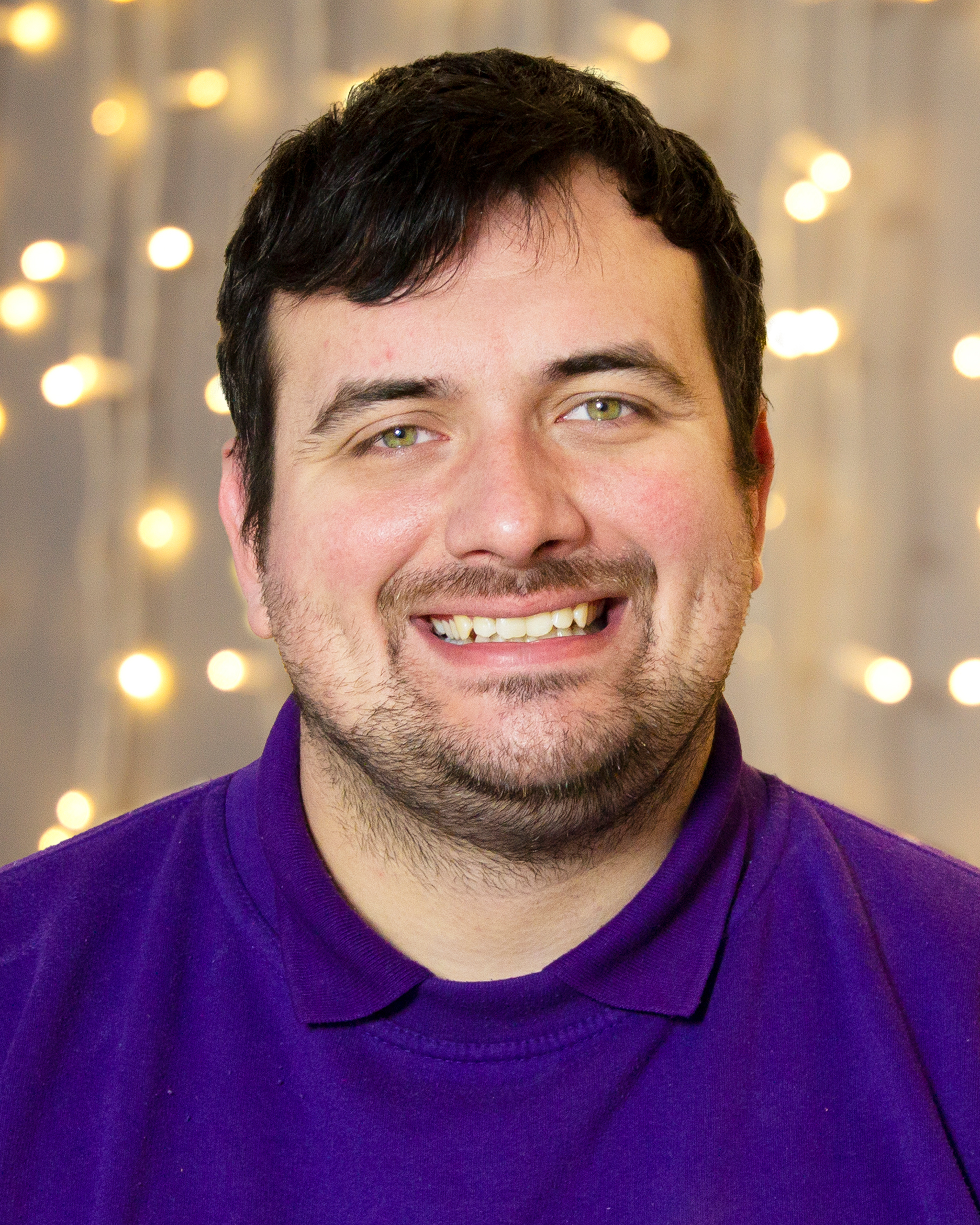 A headshot image of a Fizzco employee wearing Fizzco’s purple uniform in front of warm white curtain lights.