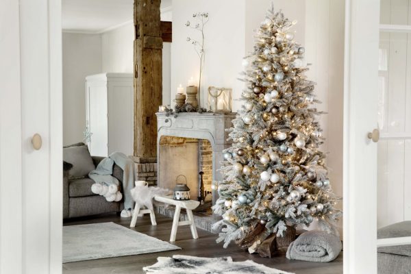 A flocked green artificial Christmas tree decorated with gold, white and blue baubles, next to a fireplace.