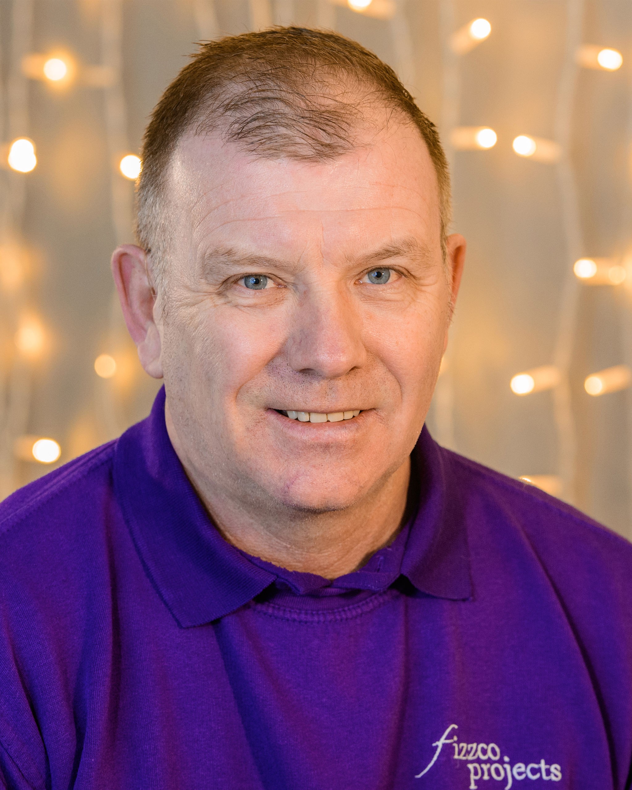 A headshot image of a Fizzco director wearing Fizzco’s purple uniform in front of warm white curtain lights.