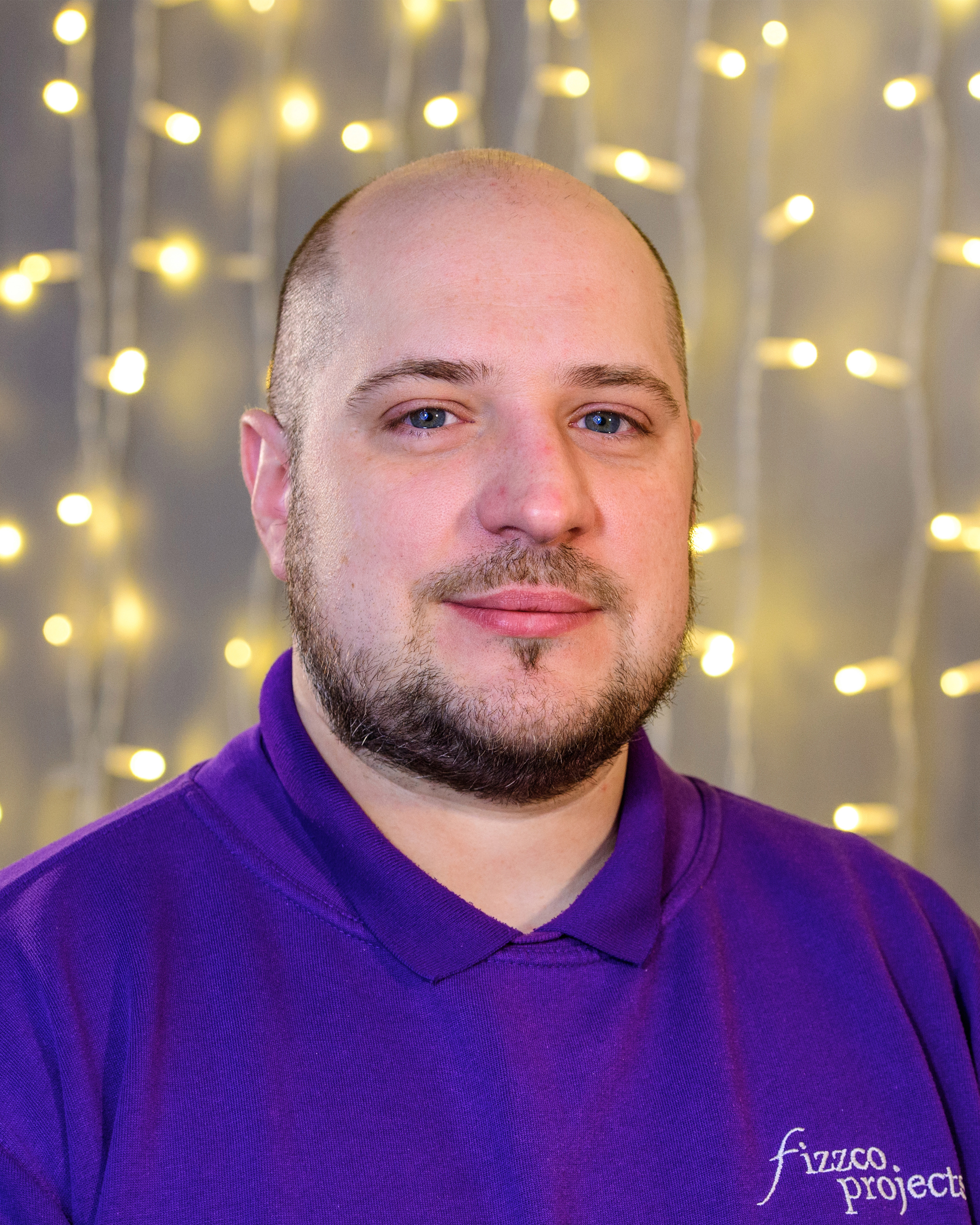 A headshot image of a Fizzco employee wearing Fizzco’s purple uniform in front of warm white curtain lights.