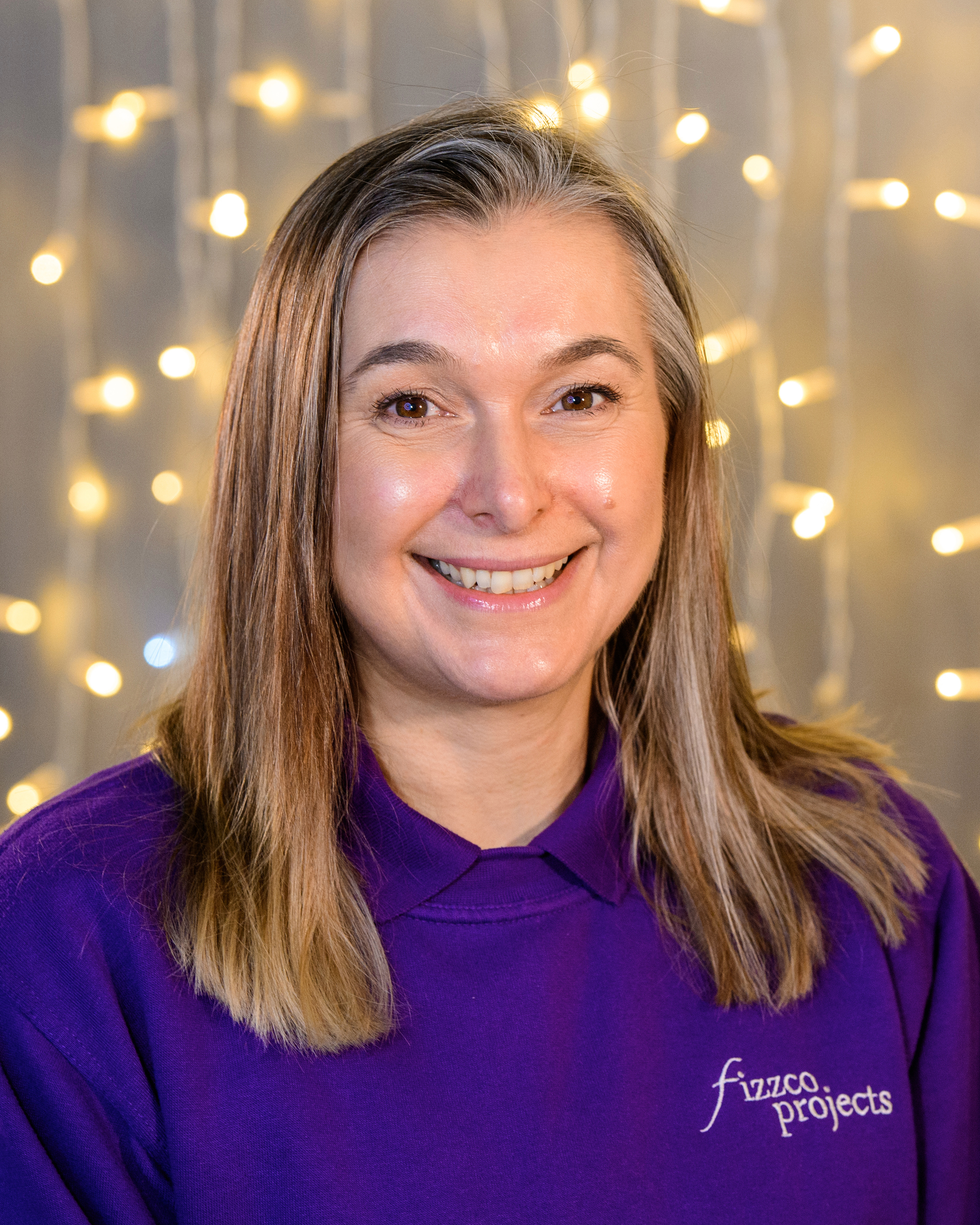 A headshot image of a Fizzco director wearing Fizzco’s purple uniform in front of warm white curtain lights.