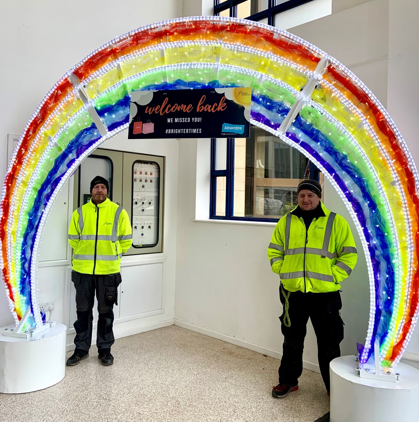 Light up rainbow arch displayed inside Alhambra Shopping Centre with two installers stood under the arch.