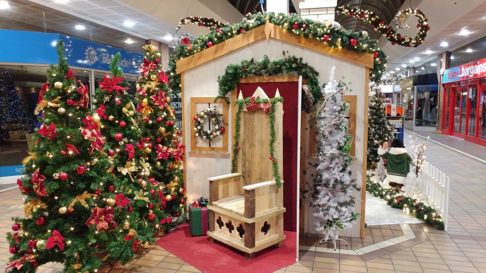A Christmas Grotto display featuring a white wooden grotto, a wooden throne and three artificial Christmas trees with red and gold baubles and flowers.