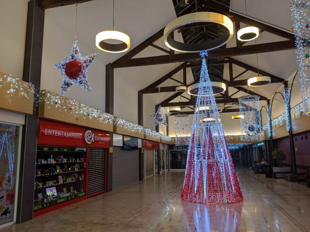 A contemporary light up red and white cone Christmas Tree in the middle of the shopping centre, with icicle lights, curtain light chandeliers and star hanging decorations all decorated with red, gold and green baubles.