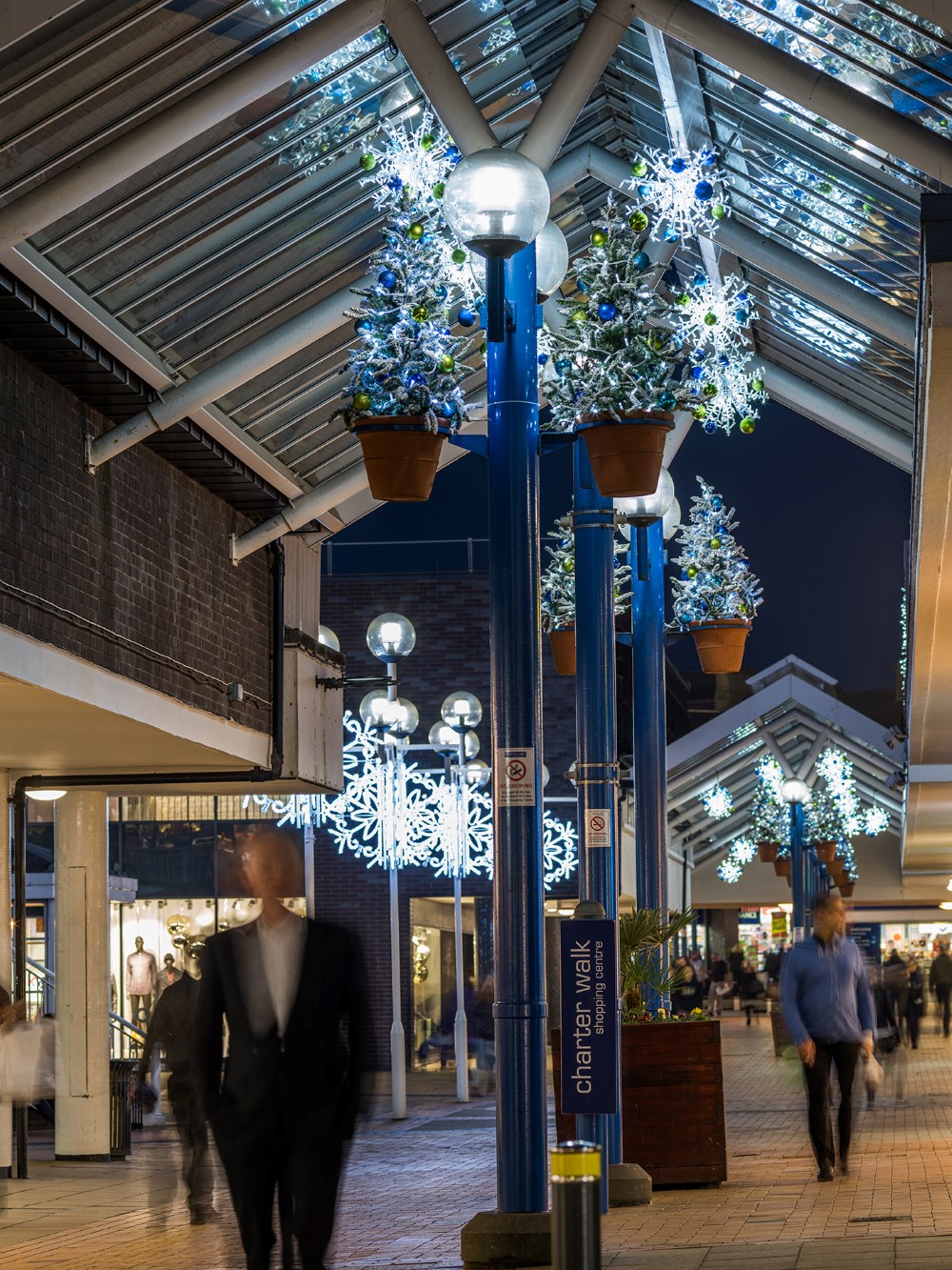 Two small Christmas trees decorated with green and blue baubles attached to each lamp post at Charter Walk Shopping Centre, and bright white snowflake motifs suspended along the walkway.