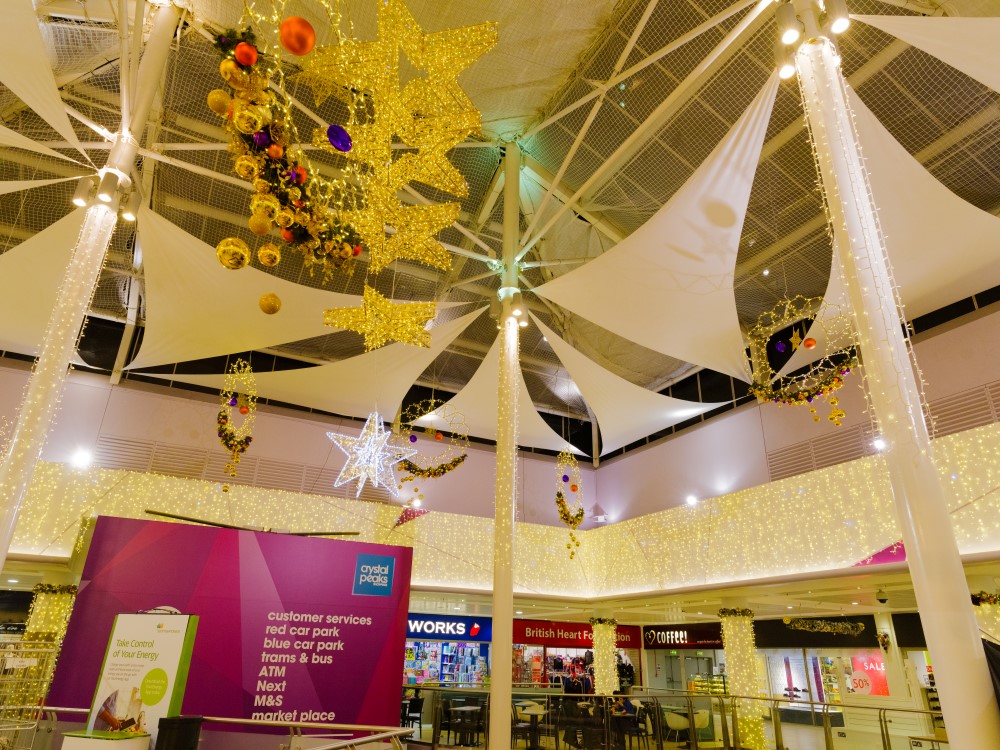 Light up gold star motifs and warm white light up silhouettes decorated with gold, copper and purple baubles suspended from the ceiling, with warm white curtain lights around the walls and pillars of Crystal Peaks Shopping Centre.