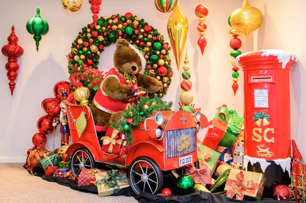A traditional Christmas Grotto display featuring a red car with a brown teddy bear driving it, with a red post box next to it and an artificial green garland decorated with red, gold and green baubles behind it, wrapped gif boxes in green, red, and gold, and large hanging decorations in red, gold and green coming down from the ceiling.