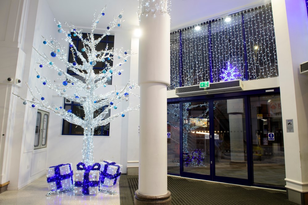 Bright white light up twig tree decorated with blue and silver baubles and light up white and blue present boxes at the base of the tree, displayed in Alhambra Shopping Centre.