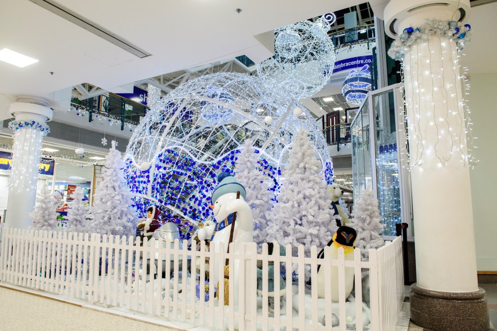A Winter Wonderland Christmas Grotto with a 3D bauble motif with bright white lights and blue tinsel and a white throne inside and a polar bear by the entrance, three plain artificial white Christmas Trees with a snowman and a penguin, and two smaller White bauble motifs hanging from the ceiling.