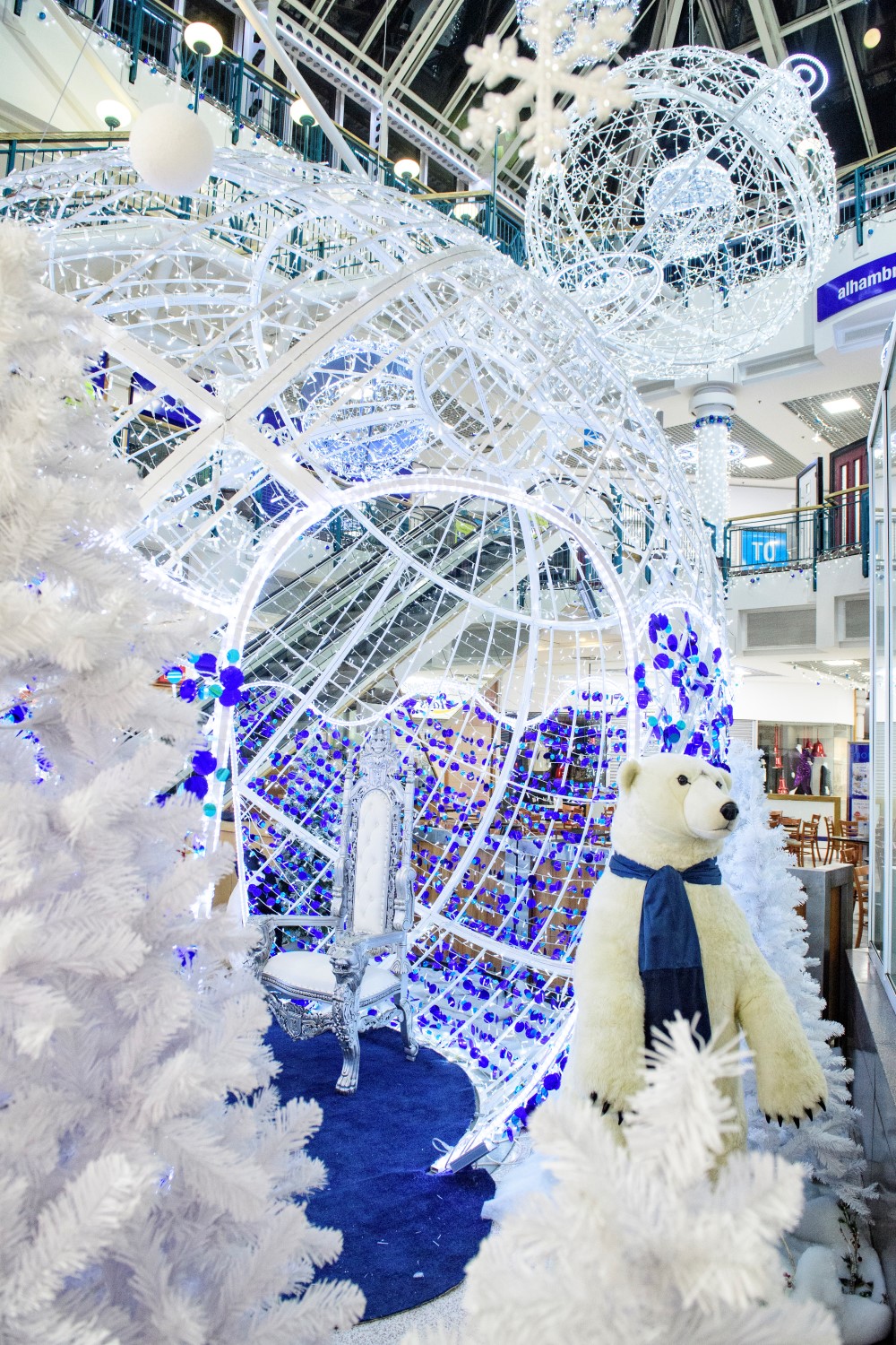 A Winter Wonderland Christmas Grotto with a 3D bauble motif with bright white lights and blue tinsel and a white throne inside and a polar bear by the entrace, three plain artificial white Christmas Trees andtwo smaller White bauble motifs hanging from the ceiling.