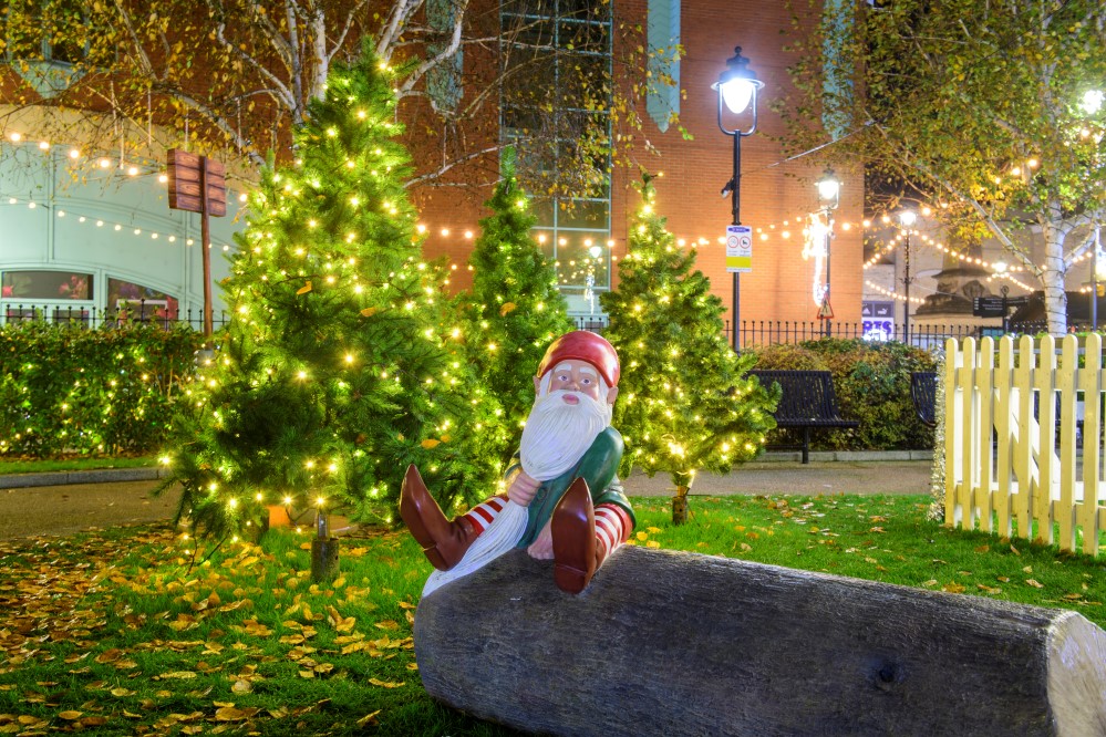 A Christmas elf sat on a log in from of three small trees decorated with warm white lights outside in St Marks, Lincoln.