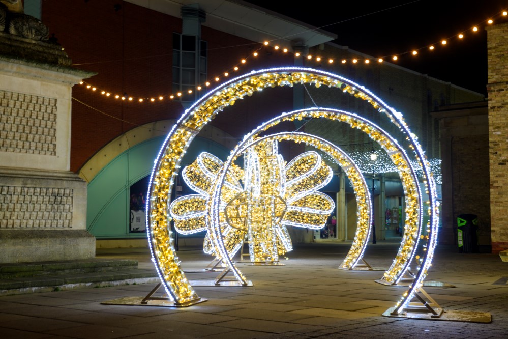 A gold and white light up 3D bow motif placed outside in the centre of St Marks, Lincoln, with warm white festoon light hanging above.