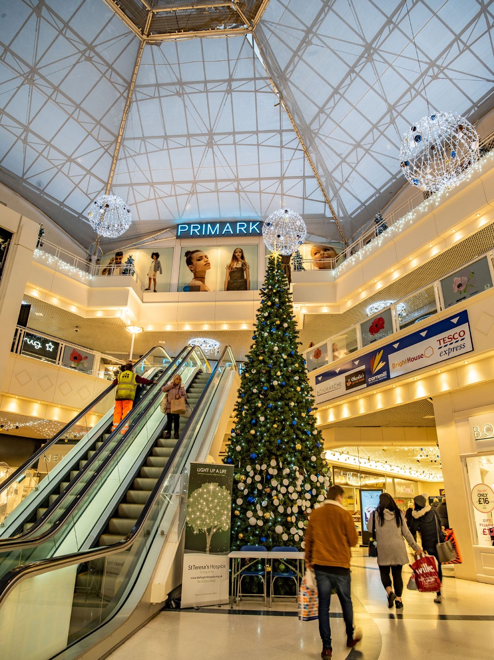 Large-scale artificial green Christmas Tree with blue, gold and white baubles on it, standing in the middle of the shopping centre with bright white icicle lights and bright white aurora balls hanging from the ceiling.