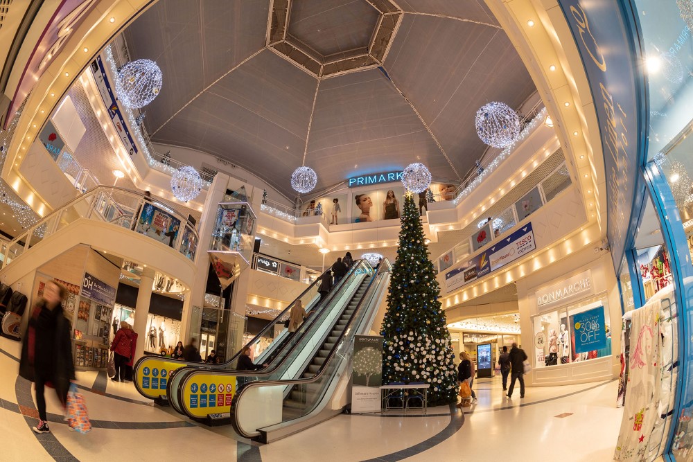 Large-scale artificial green Christmas Tree with blue, gold and white baubles on it, standing in the middle of the shopping centre with bright white icicle lights and bright white aurora balls hanging from the ceiling.