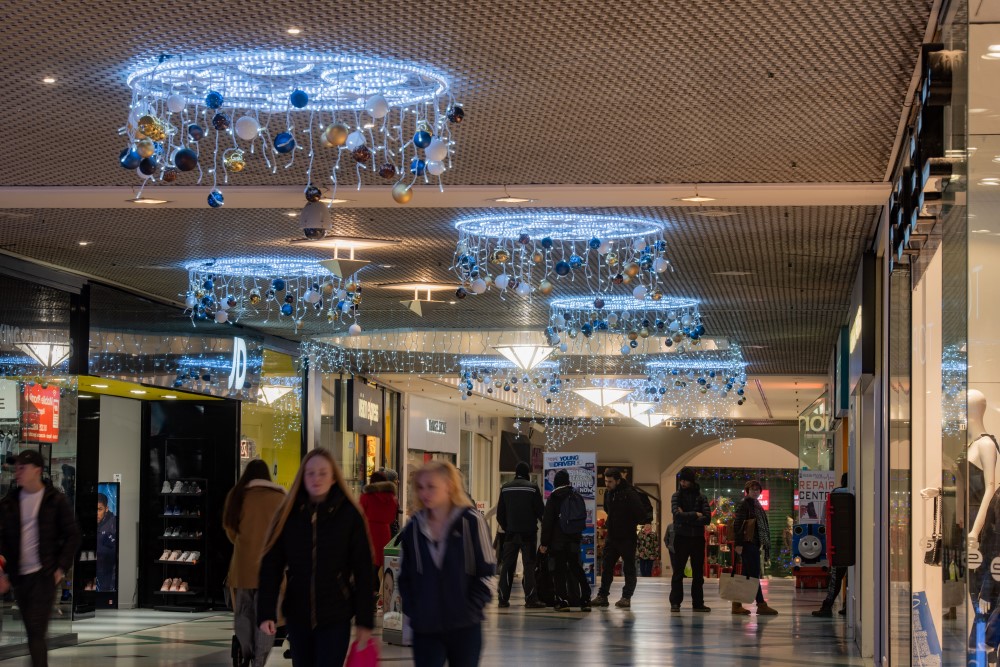 Bright white 2D circular motifs hanging from the shopping centre ceiling with blue, gold and white baubles with bright white icicle lights and bright white aurora balls hanging from the ceiling.