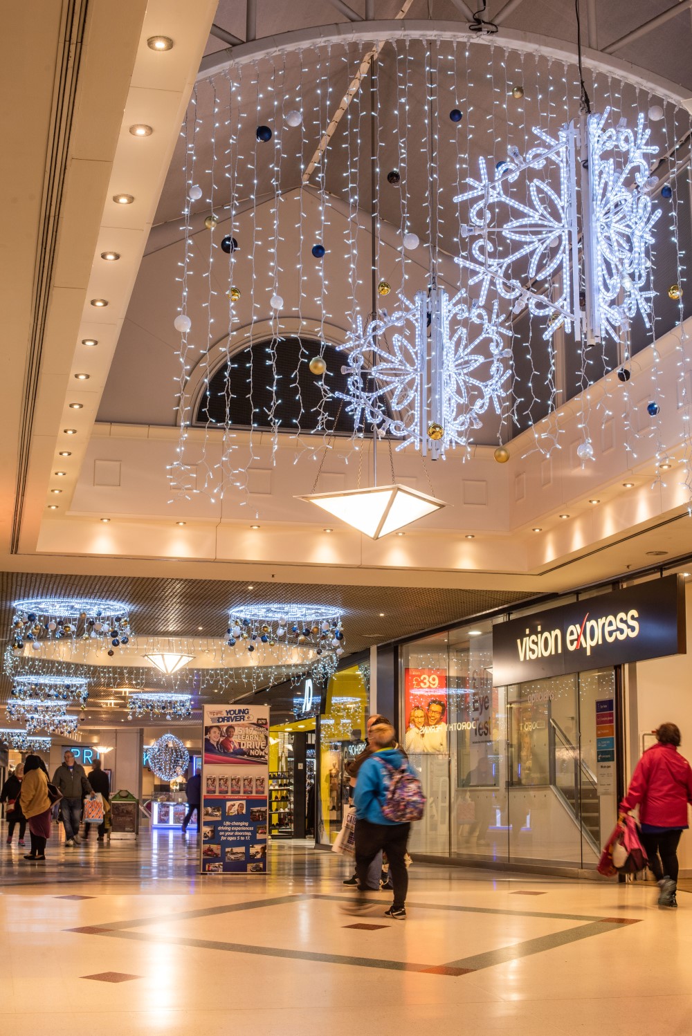 Bright white 2D circular motifs hanging from the shopping centre ceiling with blue, gold and white baubles with bright white icicle lights, curtain lights and aurora balls hanging from the ceiling.