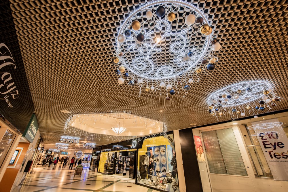 Bright white 2D circular motifs hanging from the shopping centre ceiling with blue gold and white baubles with bright white icicle lights and bright white aurora balls hanging from the ceiling.
