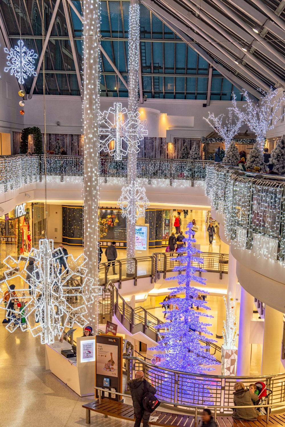 Bright white snowflake motifs suspended from the ceiling and bright white curtain lights wrapped around the pillars and the railings in the shopping centre, with a light up colour changing Christmas tree.