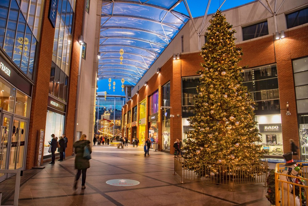 Large scale green Christmas tree decorated with gold baubles and warm white lights displayed in the centre of the shopping centre with contemporary geometric gold lights suspended from the ceiling.
