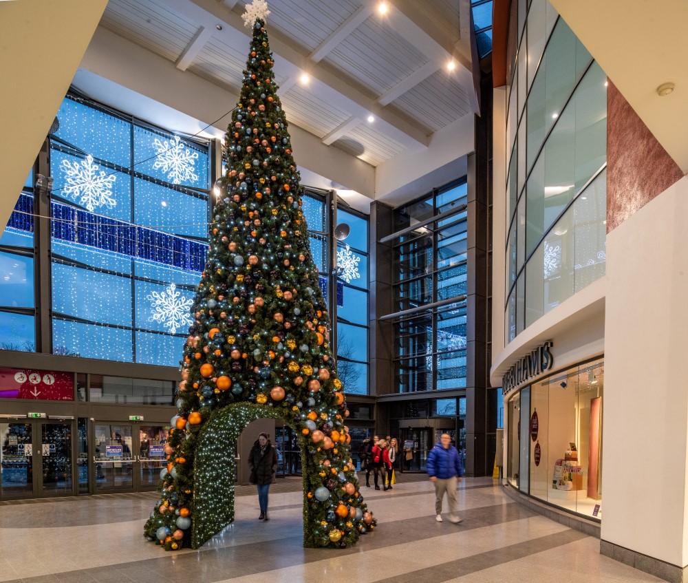 2D snowflake Motifs and curtain lights displayed on the large Shopping Centre window, a large artificial cone-shaped Christmas tree with a walk through tunnel, decorated with copper, gold and blue baubles standing in the middle of the Shopping Centre.