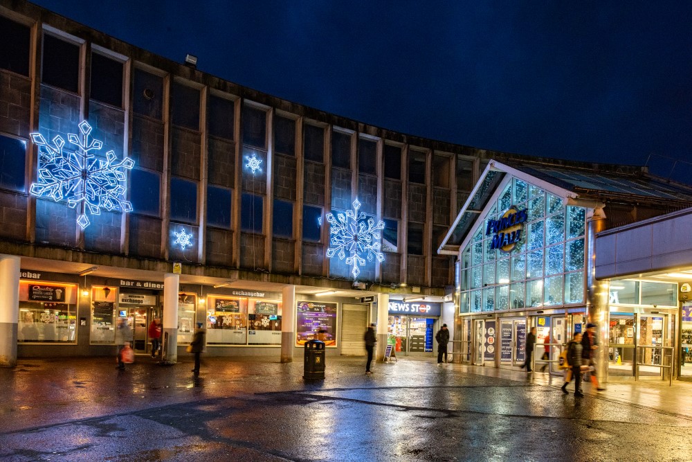 Four bright white Snowflake motifs, two large and two small, placed on the outside wall of the shopping centre and bright white curtain lights hanging across the large window above the entrance of the shopping centre.