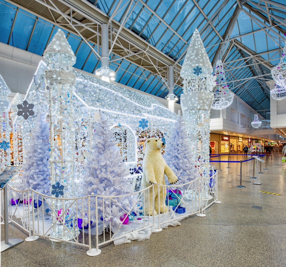 A Winter Wonderland Christmas display featuring a bright white Christmas grotto, bright white curtain lights around the pillars and bright white chandeliers decorated with light blue, dark blue and purple baubles suspended from the ceiling.