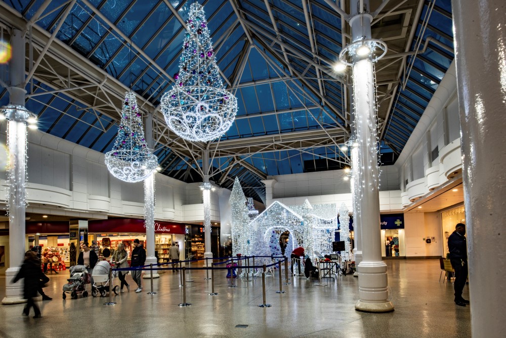 A Winter Wonderland Christmas display featuring a bright white Christmas grotto, bright white curtain lights around the pillars and bright white chandeliers decorated with light blue, dark blue and purple baubles suspended from the ceiling.