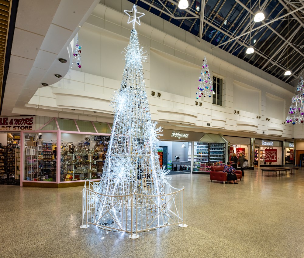 A shopping centre Christmas display featuring a bright white cone tree standing on the ground and light up spiral Christmas trees suspended from the ceiling decorated with light blue, dark blue and purple baubles.