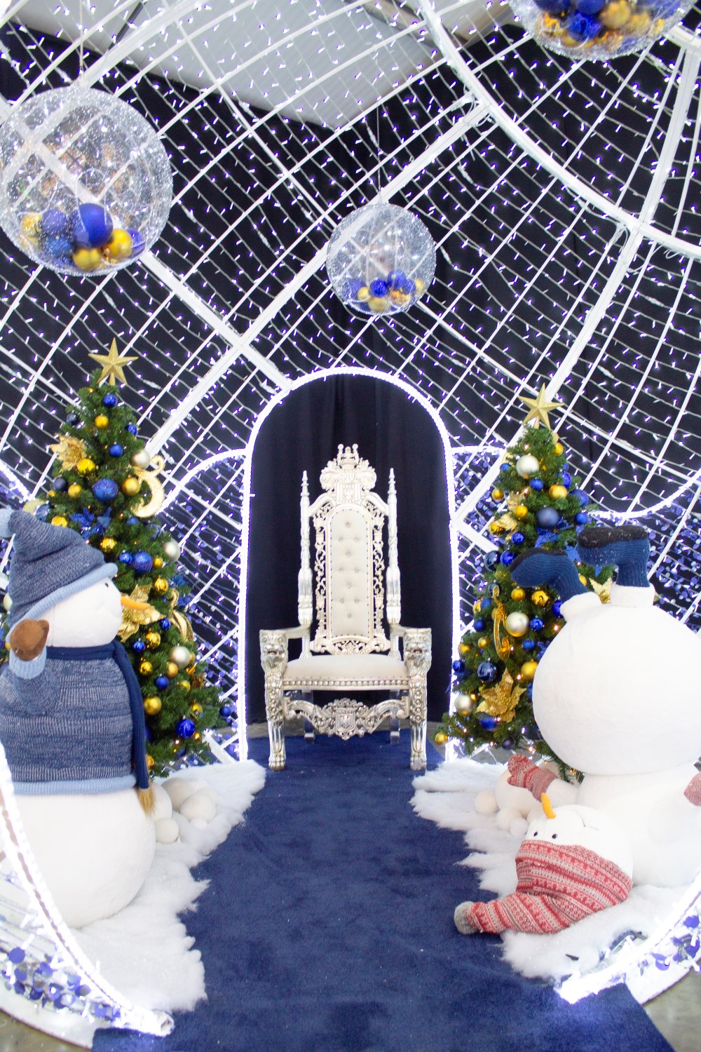 Christmas grotto scene with white throne, two blue and gold themed artificial Christmas trees, and two snowmen in a large white bauble motif with large baubles hanging down.