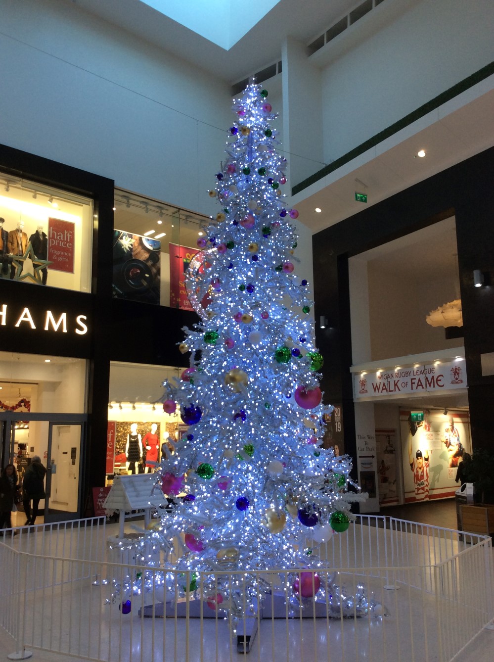 Artificial white light up Christmas Tree with pink, blue, green and white baubles attached to it, standing in the centre of a Shopping Centre.