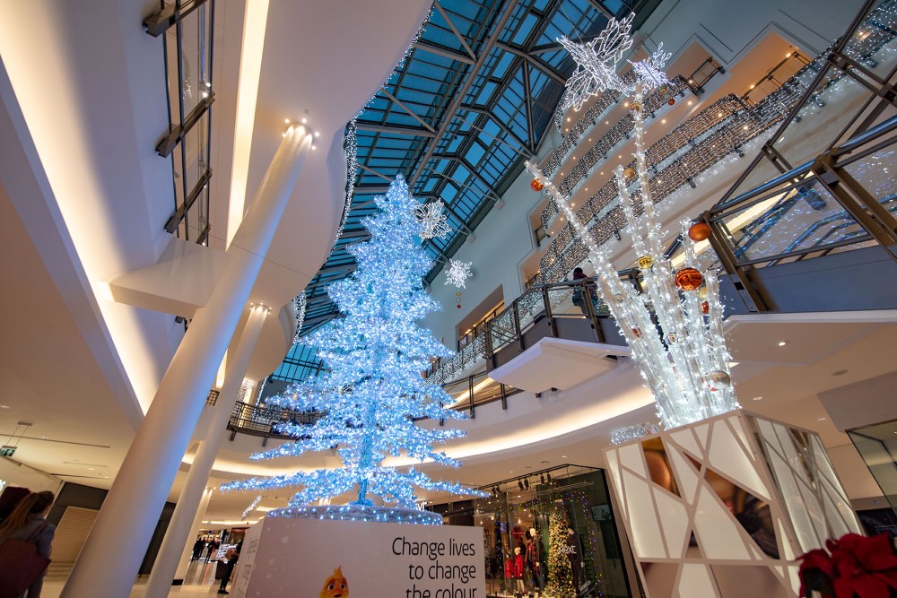 Large-scale light up colour changing artificial Christmas Tree placed in the centre of the Shopping Centre, surrounded by Bright White Curtain Lights, a light up White Vase with baubles, and a White 3D Snowflake Motif as part of a Christmas display.
