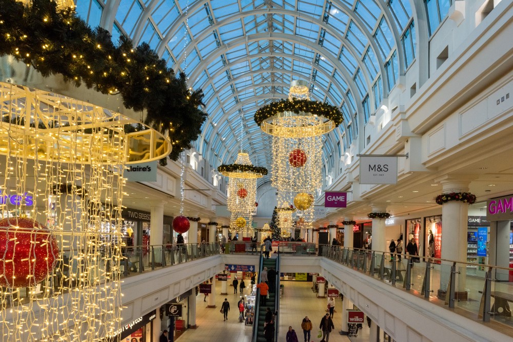 A Christmas display at Royal Priors Shopping Centre including warm white curtain light chandeliers suspended from the ceiling with green light up garlands placed around the top of them and red and gold baubles place in the centre of them.