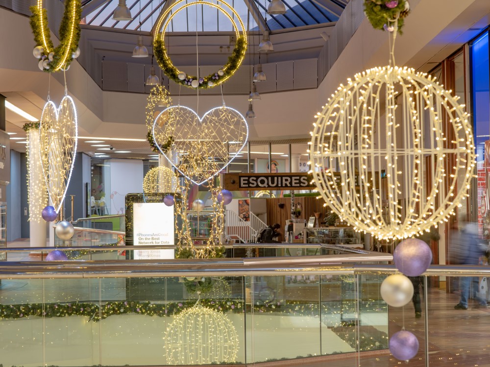 A Christmas display at the Ridings Shopping Centre featuring a large cone tree and garlands, both decorated with warm white lights white hearts and purple and silver baubles, along with warm white silhouettes in both circular and heart shapes suspended from the ceiling.