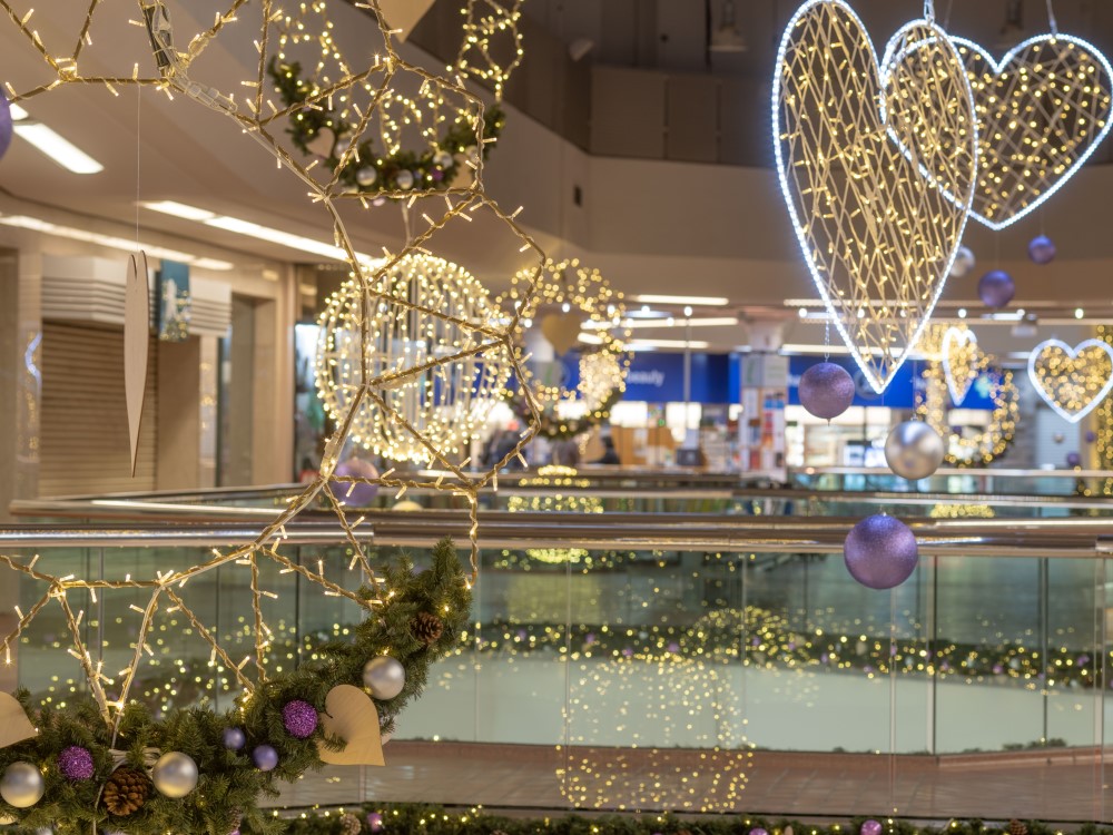 A Christmas display at the Ridings Shopping Centre featuring a large cone tree and garlands, both decorated with warm white lights white hearts and purple and silver baubles, along with warm white silhouettes in both circular and heart shapes suspended from the ceiling.
