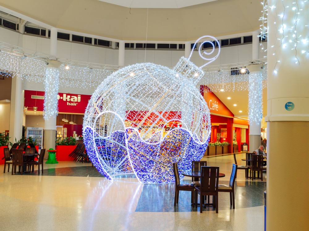 Large bright white and blue light up bauble motif displayed in the middle of the Serpentine Green Shopping Centre with bright white curtain lights suspended in the pillars around it and the ceiling above it.