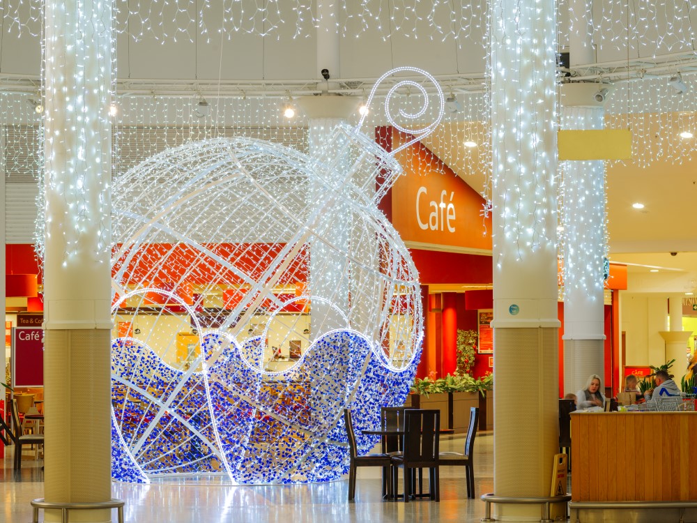 Large bright white and blue light up bauble motif displayed in the middle of the Serpentine Green Shopping Centre with bright white curtain lights suspended in the pillars around it and the ceiling above it.