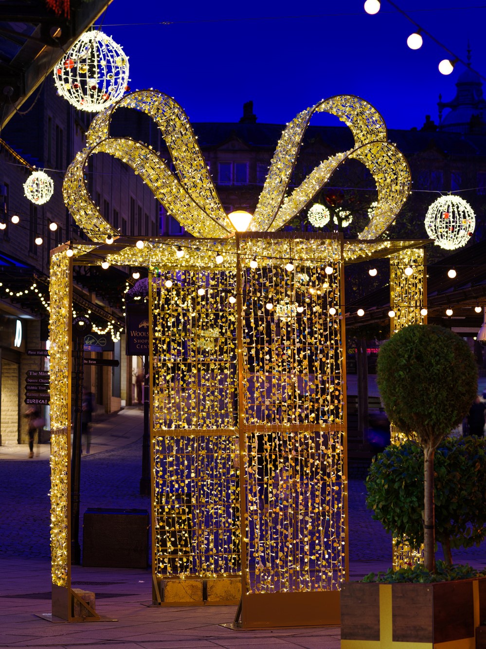Light up gold present box displayed outdoors with warm white aurora balls and festoon lights suspended above it at Woolshops Shopping Centre.