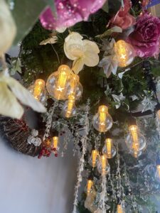 A floral arch bench featuring pink and white roses, greenery sprays, and warm white festoon lights.