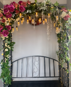 A floral arch bench featuring pink and white roses, greenery sprays, and warm white festoon lights.